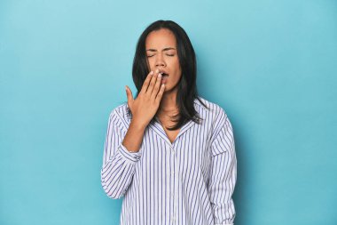 Filipinli genç bir kadın mavi stüdyoda esniyor. Ağzı eliyle kapatan yorgun bir jest gösteriyor..