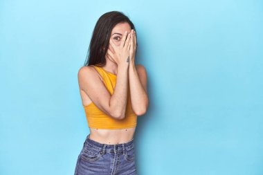 Mavi arka plandaki genç beyaz kadın korkmuş ve gergin parmaklarıyla göz kırpıyor..