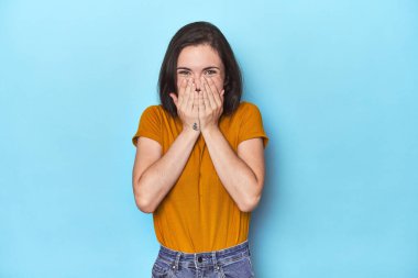 Mavi arka plandaki genç beyaz kadın bir şeye gülüyor, elleriyle ağzını kapatıyor..