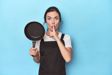 Mavi arka planda önlüğü ve tavası olan bir kadın ya sır saklıyor ya da sessizlik istiyor..