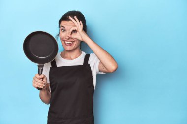 Mavi arka planda önlüğü ve tavası olan bir kadın heyecanlı..