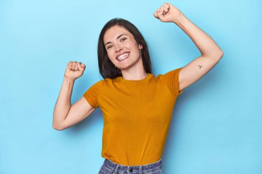 Mavi arka planda genç beyaz bir kadın özel bir günü kutluyor, atlıyor ve enerjiyle kollarını kaldırıyor..