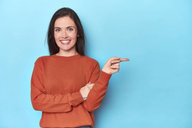 Mavi arka planda genç beyaz kadın işaret parmağıyla işaret ederek gülümsüyor..