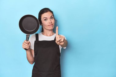 Mavi arka planda önlüğü ve tavası olan bir kadın parmaklarıyla bir numarayı gösteriyor..