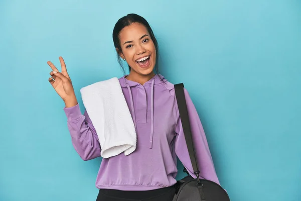 Filipina with gym gear on blue studio joyful and carefree showing a peace symbol with fingers.