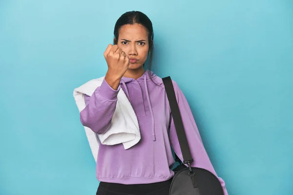 Filipina with gym gear on blue studio showing fist to camera, aggressive facial expression.