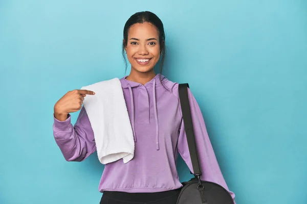 Filipina with gym gear on blue studio person pointing by hand to a shirt copy space, proud and confident