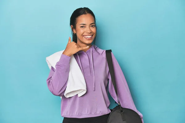 Filipina with gym gear on blue studio showing a mobile phone call gesture with fingers.