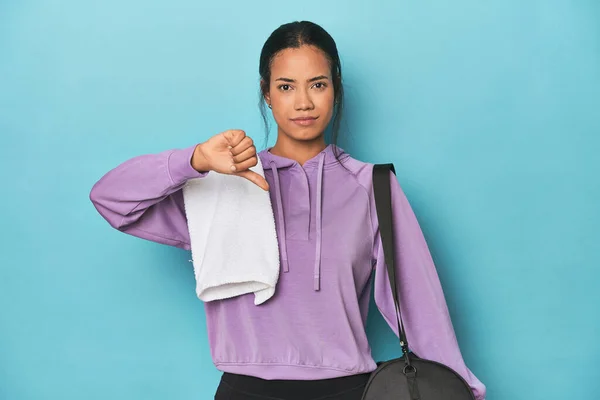 Filipina with gym gear on blue studio showing a dislike gesture, thumbs down. Disagreement concept.