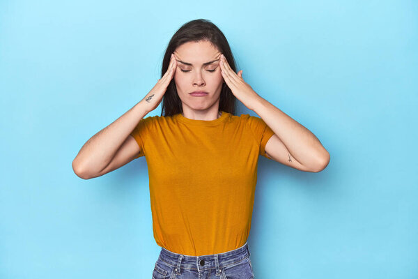 Young caucasian woman on blue backdrop touching temples and having headache.