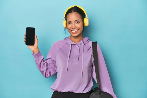 Filipina showcasing phone screen with headphones on blue backdrop