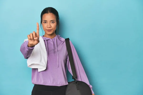 Filipina with gym gear on blue studio showing number one with finger.