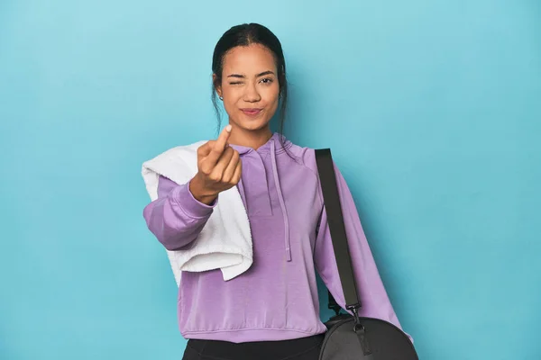 Filipina with gym gear on blue studio pointing with finger at you as if inviting come closer.