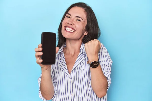 Hopeful woman displaying phone screen on blue