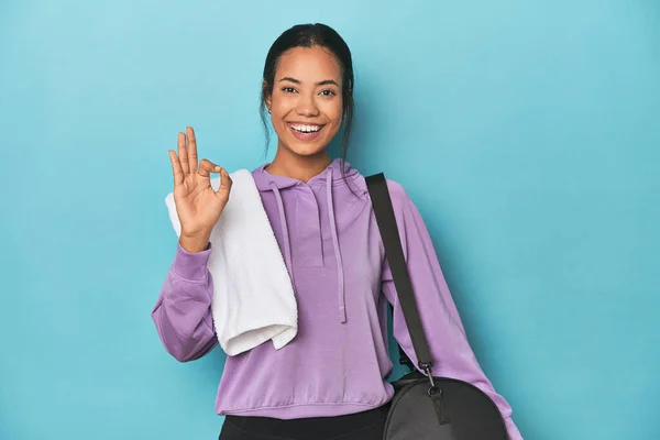 Filipina with gym gear on blue studio cheerful and confident showing ok gesture.