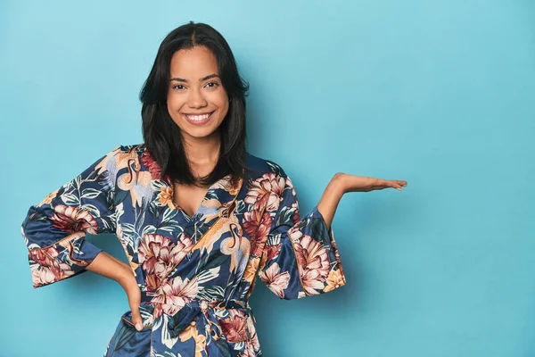 Filipina in floral kimono on blue studio showing a copy space on a palm and holding another hand on waist.