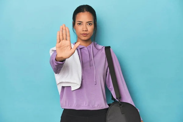 Filipina with gym gear on blue studio standing with outstretched hand showing stop sign, preventing you.