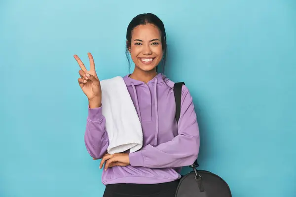 Filipina with gym gear on blue studio showing number two with fingers.