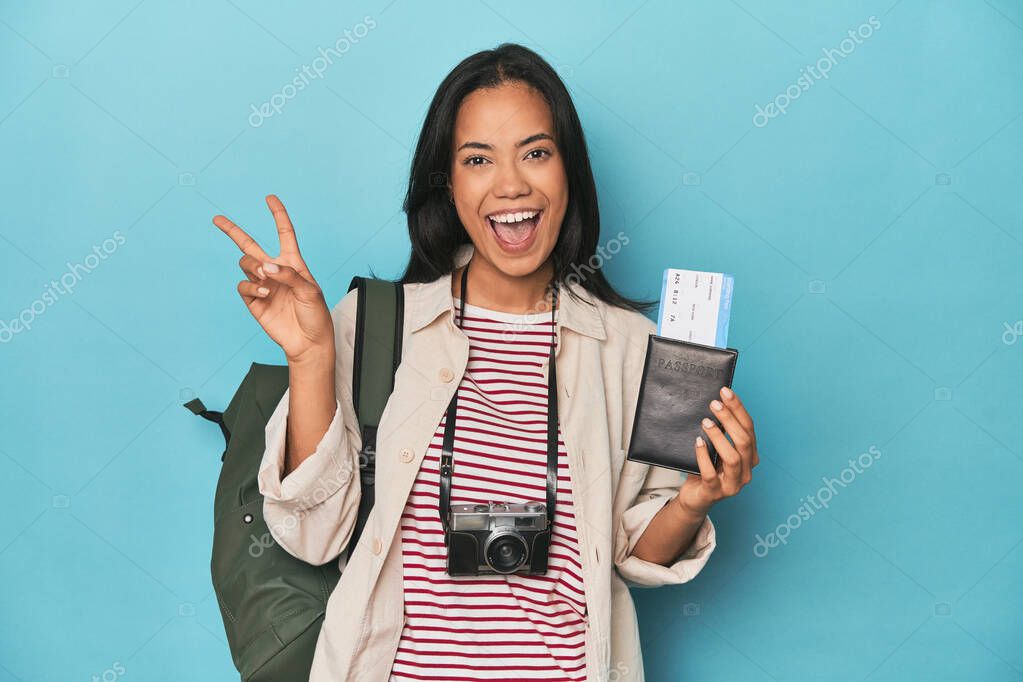 Filipina with camera, tickets, backpack on blue joyful and carefree showing a peace symbol with fingers.