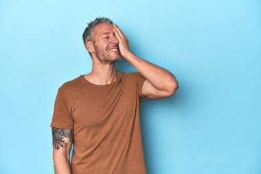 Orta yaşlı, beyaz, mavi arka planda gülen, kaygısız, doğal duygulara sahip bir adam..