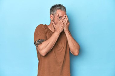 Mavi arka plandaki orta yaşlı beyaz adam korkmuş ve gergin parmaklarıyla göz kırpıyor..