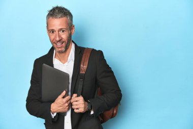 Sırt çantalı bir iş adamı dizüstü bilgisayarını mavi arkaplanda tutuyor.