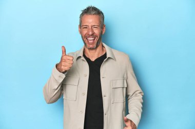 Orta yaşlı beyaz adam mavi arka planda iki parmağını da kaldırıyor, gülümsüyor ve kendinden emin..