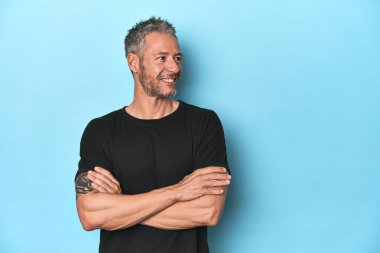 Mavi arka planda oturan orta yaşlı beyaz adam kollarını kavuşturarak gülümsüyor..