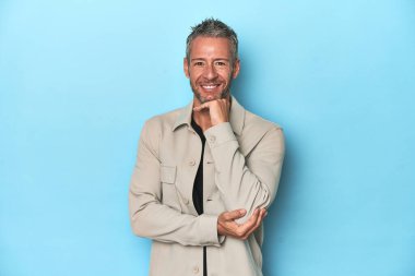 Orta yaşlı beyaz bir adam mavi arka planda mutlu ve kendinden emin gülümsüyor, çenesine elleriyle dokunuyor..