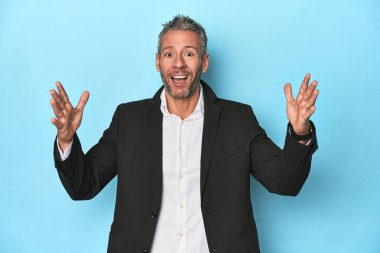 Beyaz iş adamı, mavi arka planda orta yaşlı hoş bir sürpriz alıyor, heyecanlı ve el kaldırıyor..