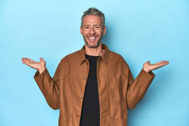 Mavi arka plandaki orta yaşlı beyaz adam kollarıyla ölçek yapıyor, mutlu ve kendinden emin hissediyor..