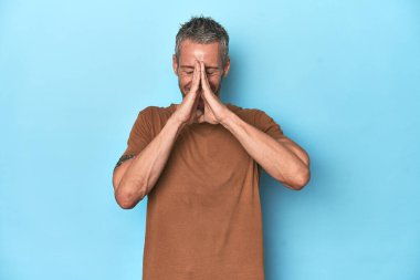 Orta yaşlı, beyaz, mavi arka planda el ele tutuşan, ağzı yakınında dua eden, kendinden emin bir adam..