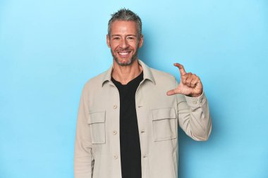 Orta yaşlı beyaz bir adam mavi arka planda. Elinde küçük bir şey tutuyor, gülümsüyor ve kendinden emin..