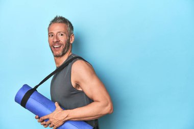 Mavi stüdyo arkaplanında paspası olan bir sporcu başparmağıyla gülüyor ve kaygısız..