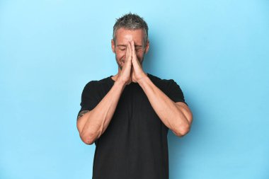 Orta yaşlı, beyaz, mavi arka planda el ele tutuşan, ağzı yakınında dua eden, kendinden emin bir adam..