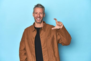 Orta yaşlı beyaz bir adam mavi arka planda. Elinde küçük bir şey tutuyor, gülümsüyor ve kendinden emin..