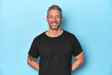 Mavi arka plandaki orta yaşlı beyaz adam mutlu, güler yüzlü ve neşeli..