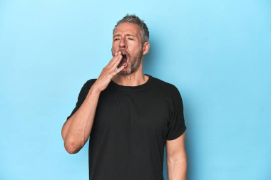 Orta yaşlı beyaz adam mavi arka planda esniyor. Ağzı eliyle kapatan yorgun bir hareket sergiliyor..