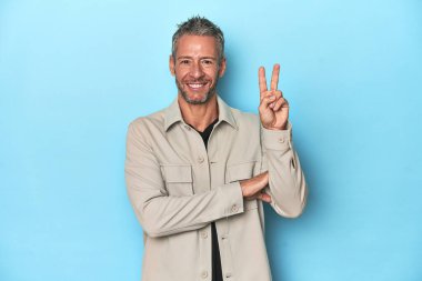 Mavi arka planda orta yaşlı beyaz bir adam iki numarayı parmaklarıyla gösteriyor..
