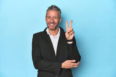 Beyaz iş adamı, orta yaşlı, mavi arka planda iki numarayı parmaklarıyla gösteriyor..