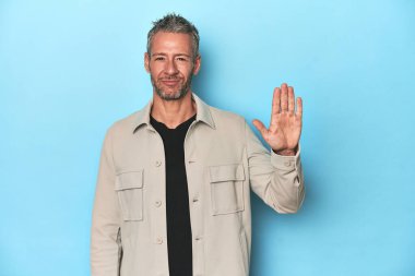 Mavi arka planda orta yaşlı beyaz bir adam gülümseyerek beş numarayı parmaklarıyla gösteriyor..