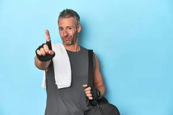 Athlete with gym backpack on blue background showing number one with finger.