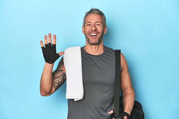 Athlete with gym backpack on blue background smiling cheerful showing number five with fingers.