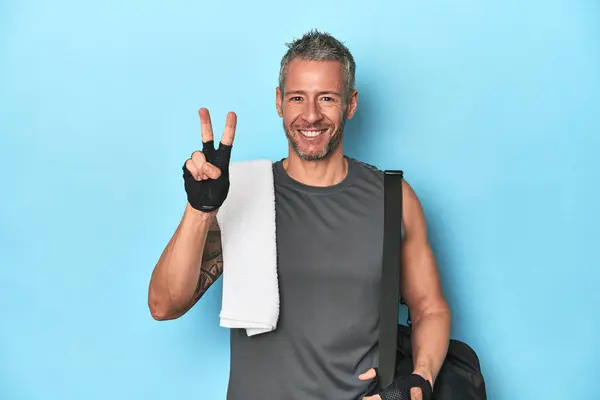 Athlete with gym backpack on blue background showing number two with fingers.
