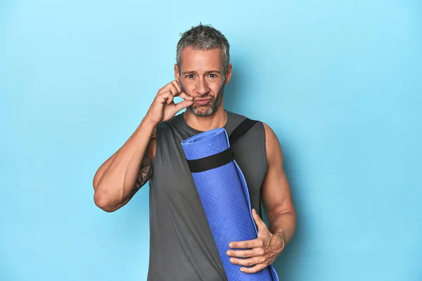 Athlete with a mat on a blue studio backdrop with fingers on lips keeping a secret.