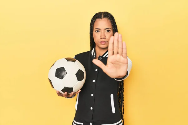 Indonesian schoolgirl with soccer ball on yellow standing with outstretched hand showing stop sign, preventing you.