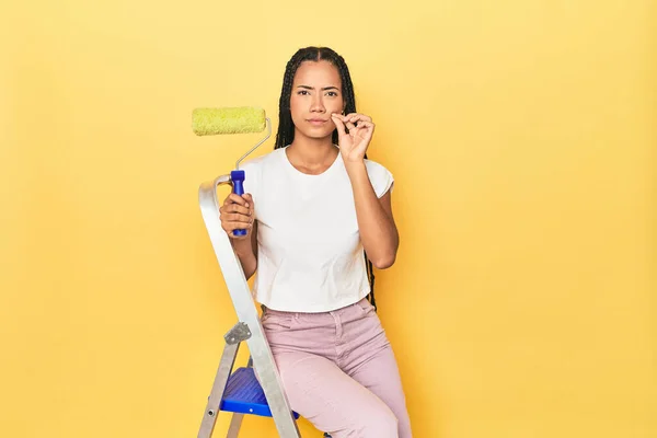 Indonesian woman with roller on ladder on yellow with fingers on lips keeping a secret.