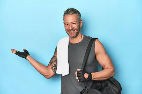 Athlete with gym backpack on blue background showing a copy space on a palm and holding another hand on waist.