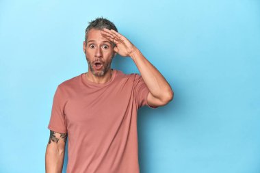 Mavi arka plandaki orta yaşlı beyaz adam yüksek sesle bağırıyor, gözleri açık ve elleri gergin tutuyor..