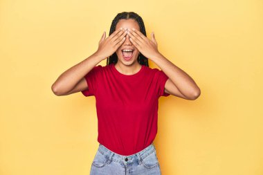 Sarı stüdyo arkaplanındaki Endonezyalı genç kadın elleriyle gözlerini kapatıyor, geniş bir gülümsemeyle bir sürpriz bekliyor..